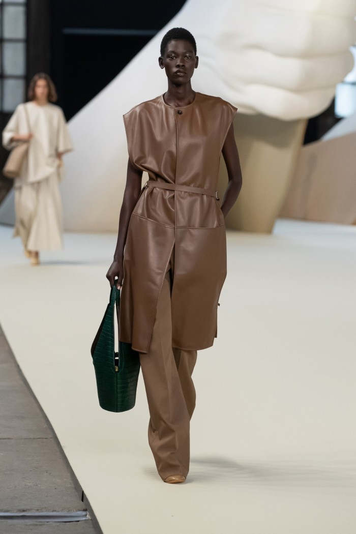 A female catwalk model in long sleeveless leather dress in cocoa brown, worn over trousers of a similar colour and carrying a black handbag