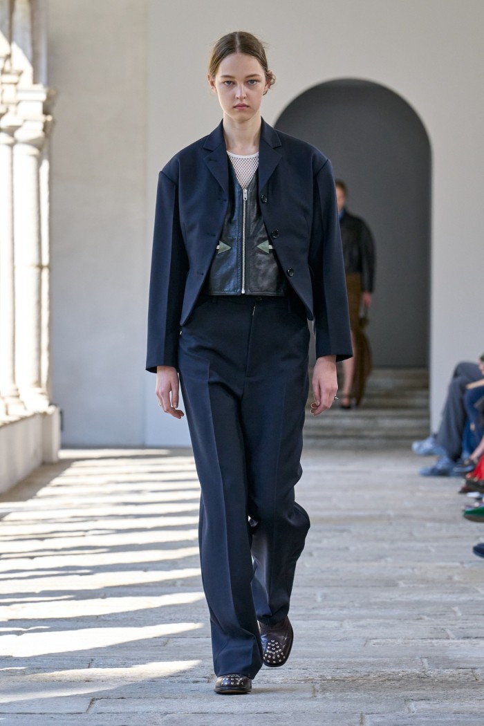 A female catwalk model in a dark-blue trouser suit of short tailored jacket and high-waisted trousers with a leather-look waistcoat