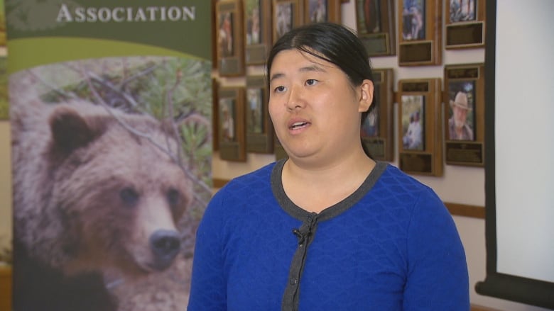 a person in a blue sweater stands in front of a wall of plaques and photographs. there is a banner with a picture of a grizzly bear.