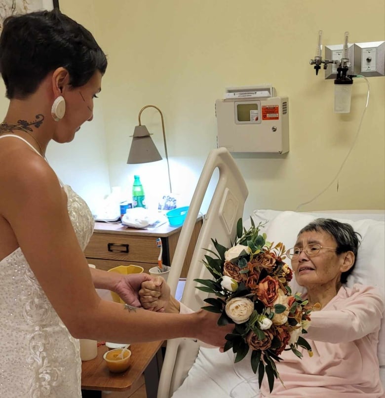 A woman in a wedding dress stands next to a woman in a hospital bed wearing pink pajamas. They were holding hands.