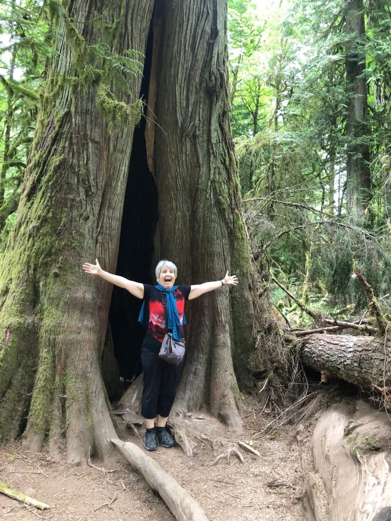 A white-haired woman stands in front of a giant tree, with her arms spread wide. 