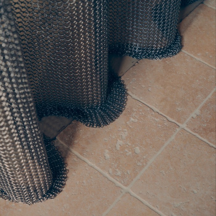 The bottom edge of a custom designed stainless steel chainmail shower curtain drapes on a tiled floor