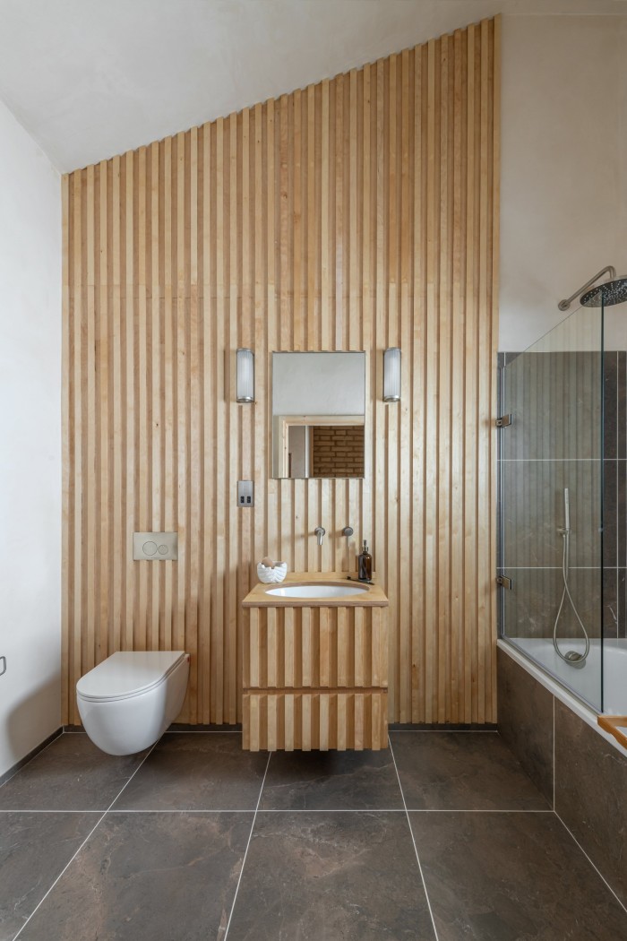 A bathroom, featuring a sink and toilet against a wood-panelled wall. The cupboard under the sink is also wood-panelled to match. To the right, there is a bath, with a glass shower wall. The floor is tiled in dark grey