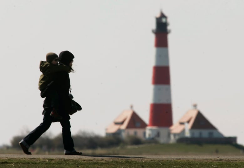 A woman carries a child on her back in front of a lighthouse