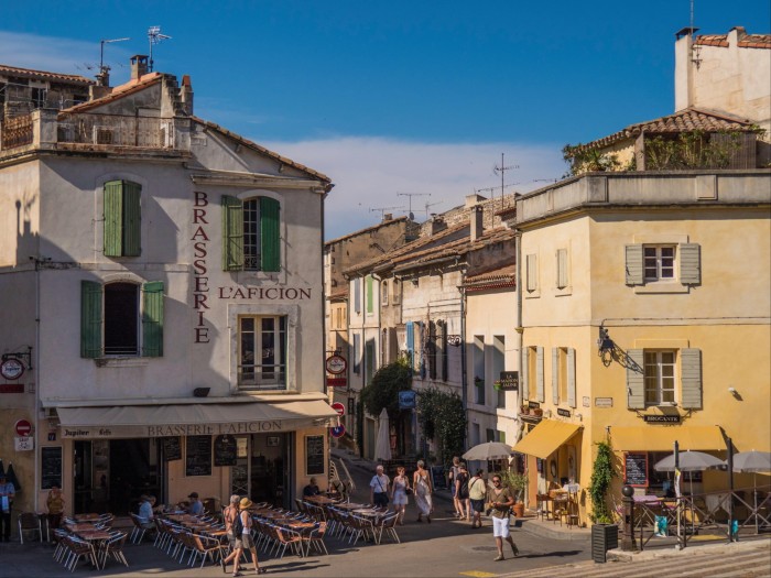 A street in a southern French town featuring traditional buildings with wooden shutters and people sitting in the sun or under parasols outside cafés