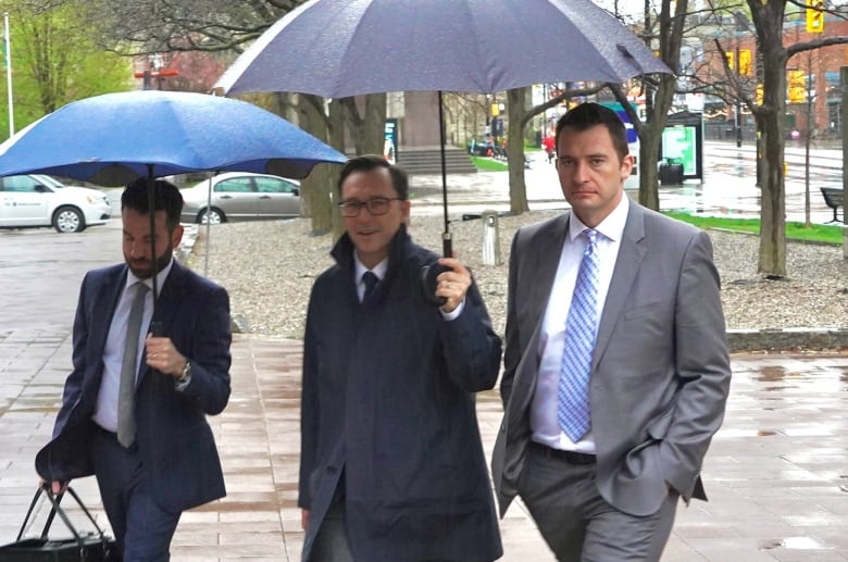 Three men in suits and ties, two of whom have umbrellas, walk outside on a cloudy day.