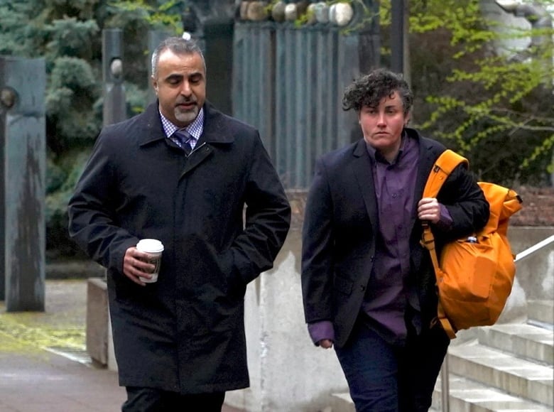 Two people — one carrying an orange bag, the other holding a coffee cup — walk down a city street on a cloudy day.