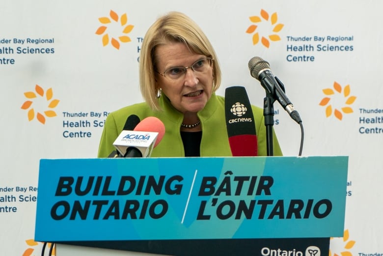 A woman with a blond bob and glasses leans into microphones on a podium in front of a backdrop that reads "Thunder Bay Regional Health Sciences Centre". 