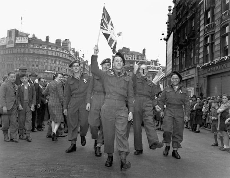 Canadian soldiers waving a British flag march in formation through the middle of a city with smiles on their faces as an appreciated crowd lines the streets.