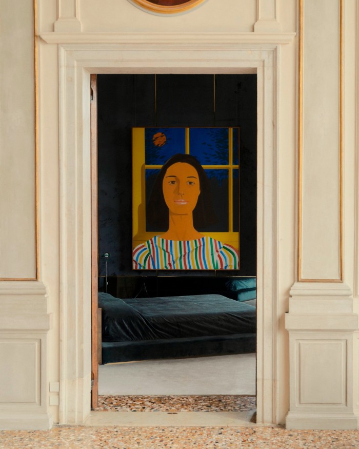 A brightly coloured painted portrait of a woman in a stripy top is hung on a dark-blue wall, seen through an ornate, gilt-edged door frame 