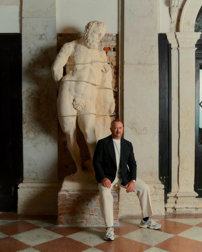 A man in trainers, cream-coloured trousers and a black jacket stands in a room with a chequerboard marble floor, next to a towering ancient male figurative sculpture 