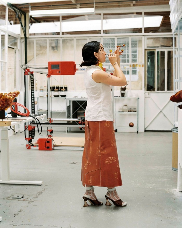 A young women in a sleeveless white top, a long, terracotta-coloured skirt and heeled mules stands in the middle of a room with glass-partitioned walls. She is blowing into a metal tube, creating an amber-coloured bubble 