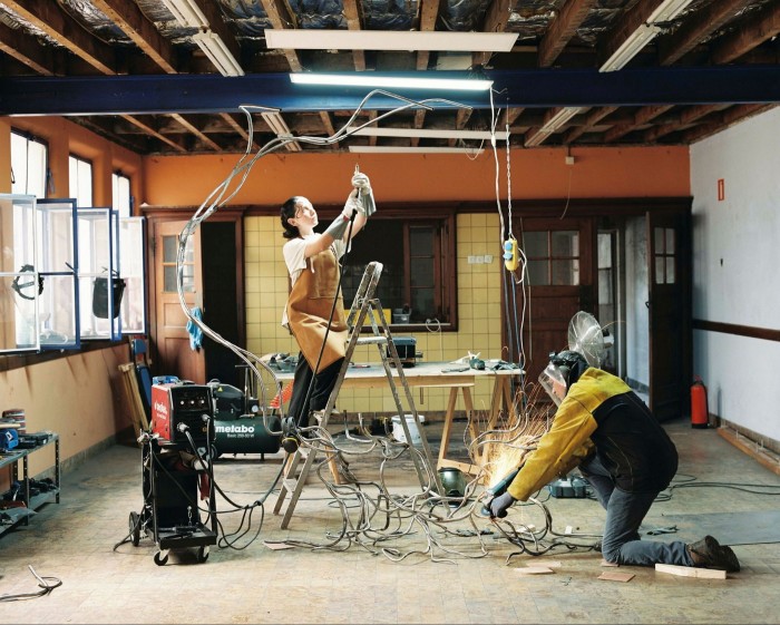 A young women stands on a step ladder wearing a brown apron in the middle of room with a bare-beam ceiling, and orange and yellow walls. She is surrounded by machines and a ceiling sculpture of tangled steel branches. A man works on the sculpture with an angle grinder 