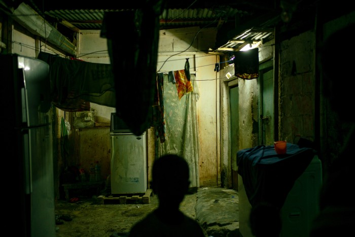 A dimly lit interior of a run-down home, with hanging clothes, an old refrigerator, and makeshift curtains