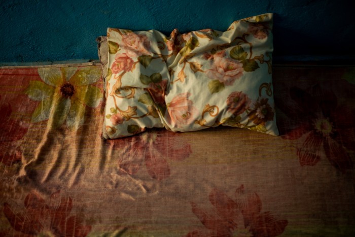 Covered in an almost coordinating pink floral fabric, a pillow sits on a dirty, uncovered mattress propped up against the wall of a blue rented room