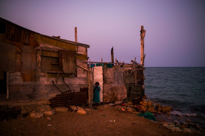 A young child standing near the entrance of a makeshift wooden shack by the ocean at dusk