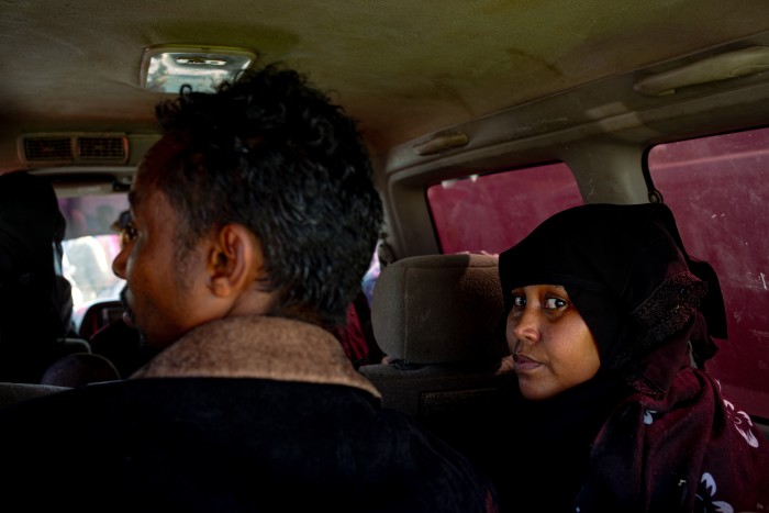 A woman in a black headscarf sitting in the backseat of a vehicle, looking towards the camera, while a man seated in front of her faces forward