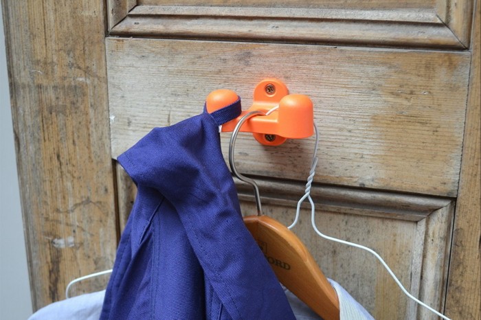 The bright-orange double hook is shown in close-up on a well-worn wooden door, holding a work jacket in primary blue and a white shirt on a coat hanger 