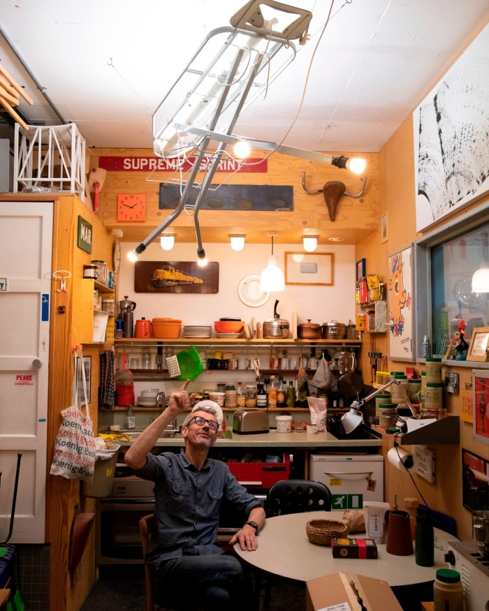 A man wearing black-rimmed glasses, a blue shirt and jeans sits at table in a kitchen with yellow walls, pointing at a ceiling light made out of an ironing-board frame