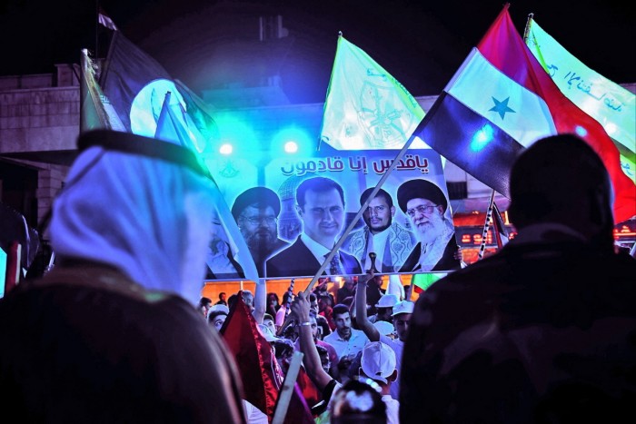 Syrians wave flags and lift a placard depicting Hassan Nasrallah, Syrian President Bashar al-Assad, Yemen’s Houthi leader Abdulmalik al-Houthi, and Iran’s Supreme Leader Ayatollah Ali Khamenei, at a rally in 2021