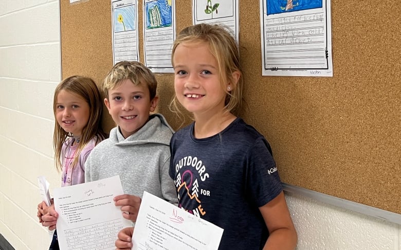 Three smiling elementary students stand in a row in a school hallway, each showing off a written assignment about local plants. 