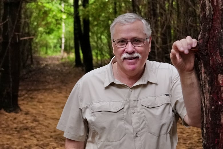 A grey-haired man in a short sleeve shirt stands smiling in a forest clearing, with his hand resting against a tree.