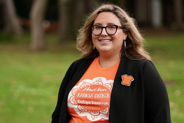 A woman in an orange t-shirt and black cardigan, with a beaded orange t-shirt broach pinned to it, stands smiling in a park setting.