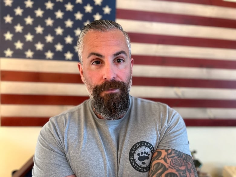 A muscular man with a goatee, wearing a grey T-shirt, sits in front of an American flag. 