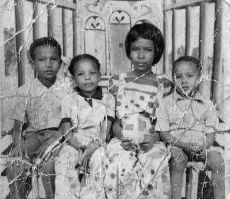 A black and white photograph, full of scratches as if it's been folded or crumpled, shows four young Sudanese children sitting side by side.