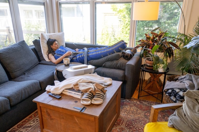 A woman is lying on a grey sectional couch. She is in a blue suit that looks like a blanket. 