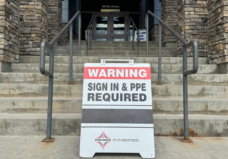 A sandwich board sign bearing the message 'Warning: sign in & PPE required' stands outside the front steps of a school entrance.