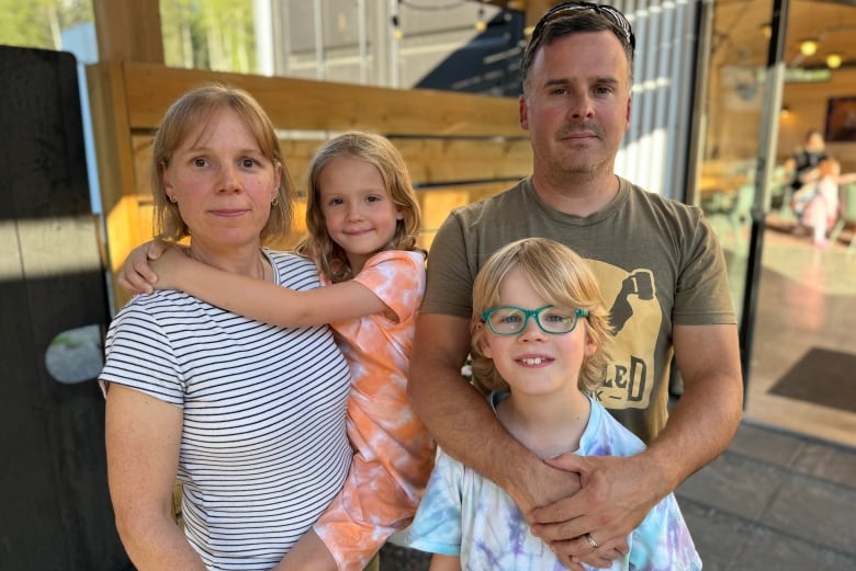 A family poses outside a restaurant: a woman carries her young daughter while standing next to her husband, who clasps his arms around their son's shoulders in front of him.