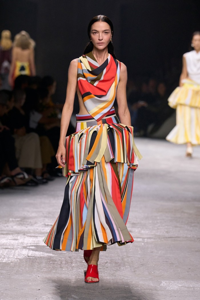 A female catwalk model wearing a long, wide-skirted sleeveless dress in stripes and tassels of many colours