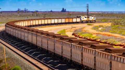 A long freight train carrying iron ore travels along a rail track on a clear day, approaching Port Hedland, Australia. The landscape around the railway is arid, with patches of sparse vegetation and reddish soil. In the background, industrial structures are visible.