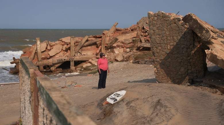 A woman in a pink shirt stands on the beach next to a crumbled stone wall, with rubble in the background extending into the water. 