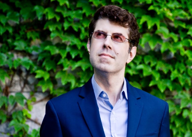 A man in a dark blue suit and button-down shirt looks up to the left as he stands outside near an ivy covered rockface.