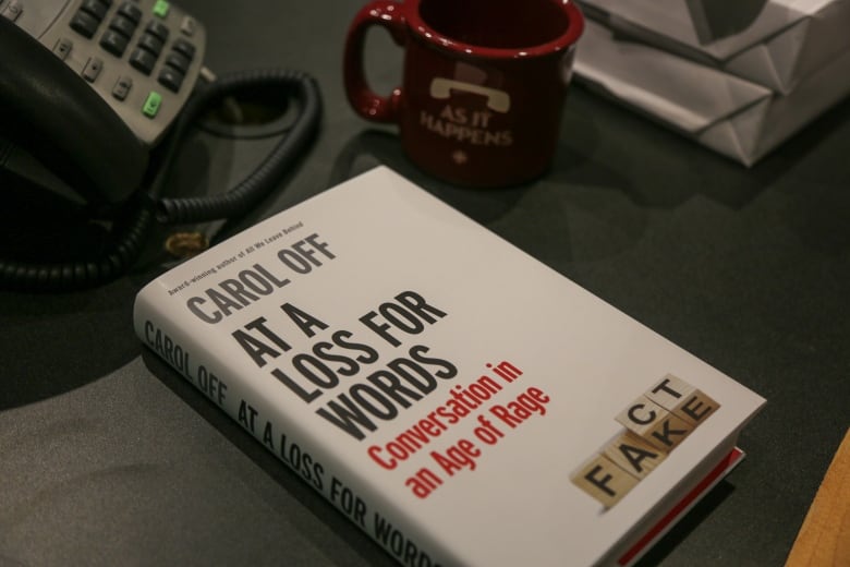 A hardcover book on a desk next to a phone and an "As It Happens" branded mug. The title, in black text on white background, reads: "At A Loss For Words: Conversation in an Age of Rage." In the bottom right corner are four scrabble tiles, with the last two turned to reveal extra letters, simultaneously spelling "Fact" and "Fake."