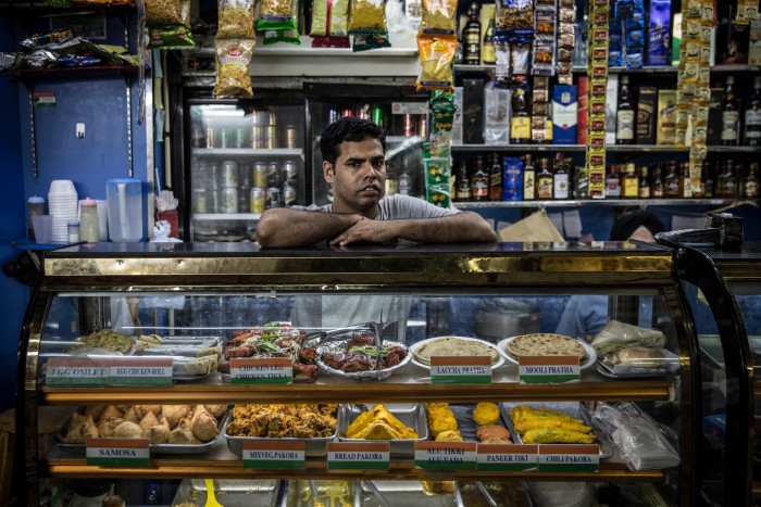 An Indian man standing behind a glass display case that contains a variety of Indian food