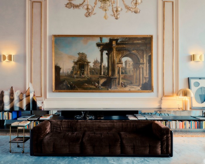 The large and dramatically lit painting is surrounded by, gilt wall mouldings, an ornate glass chandelier and a modern, boxy brown velvet sofa 