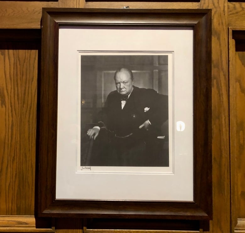 A framed black and white photo of a scowling man is shown on a wood-panelled wall.