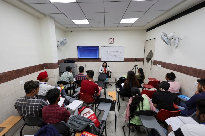 An instructor sits at the front of a room while students sit listening.