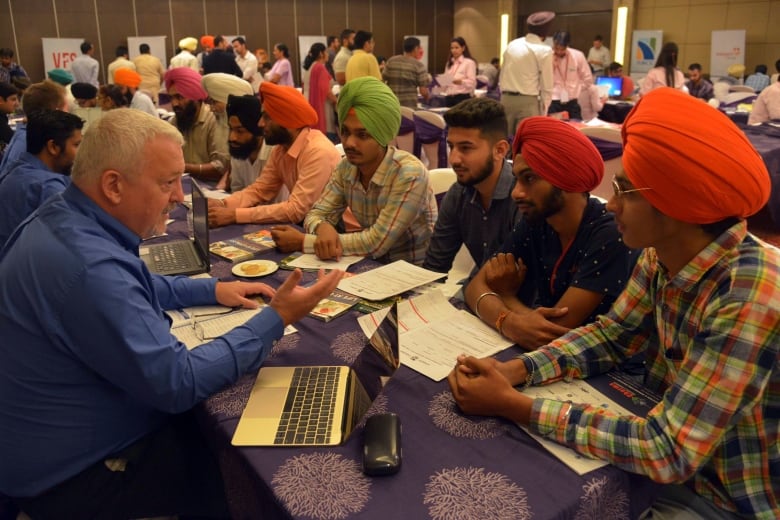 An adult sitting on one side of a table talks to several younger adults sitting on the other side of the table.