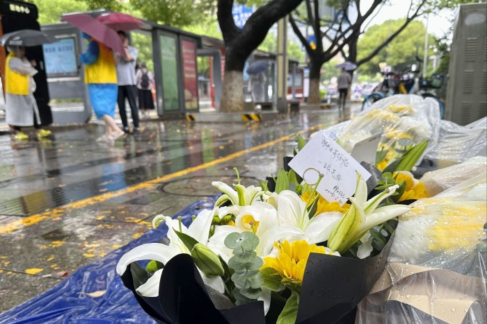 Bouquets of flowers, including white lilies and yellow daisies, are placed on the ground near a bus stop in Suzhou. In the background, people holding umbrellas are seen standing and walking on a rainy day.