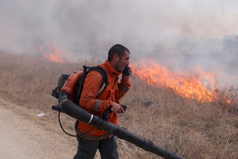 A man with significant stubble talks on a mobile device while carrying a hose-like device. Behind him, orange flames are shown in the brush.