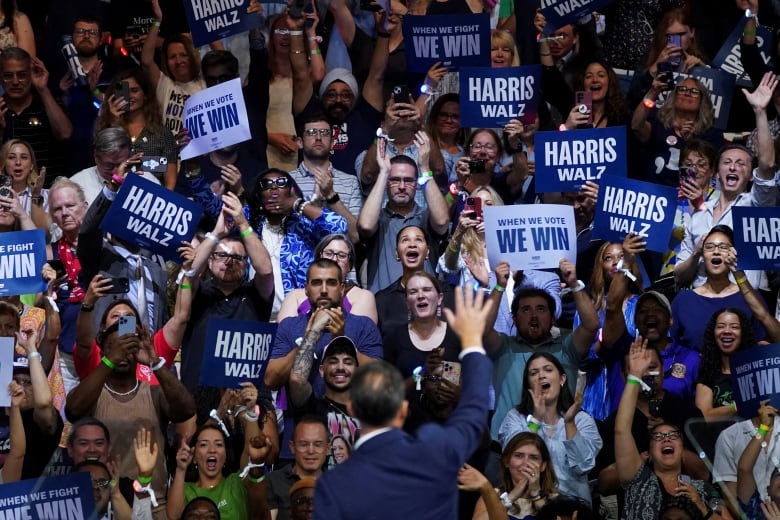 A man is shown in the foreground with his back to the camera waving his right arm in the air from stage, in front of a large crowd holding signs that say 'Harris-Walz.'