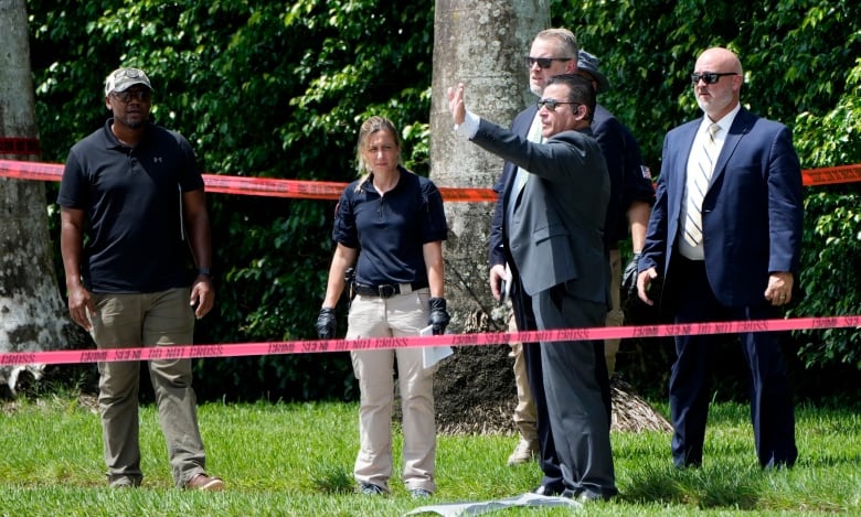 A group of law enforcement individuals stand in front of red police tape around a line of trees in the background. 