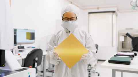 A person in a cleanroom suit, mask, and glasses holds a large, square, golden-colored chip, Cerebras’s CS-3 chip, in a laboratory setting. In the background, there are computer monitors and various lab equipment.