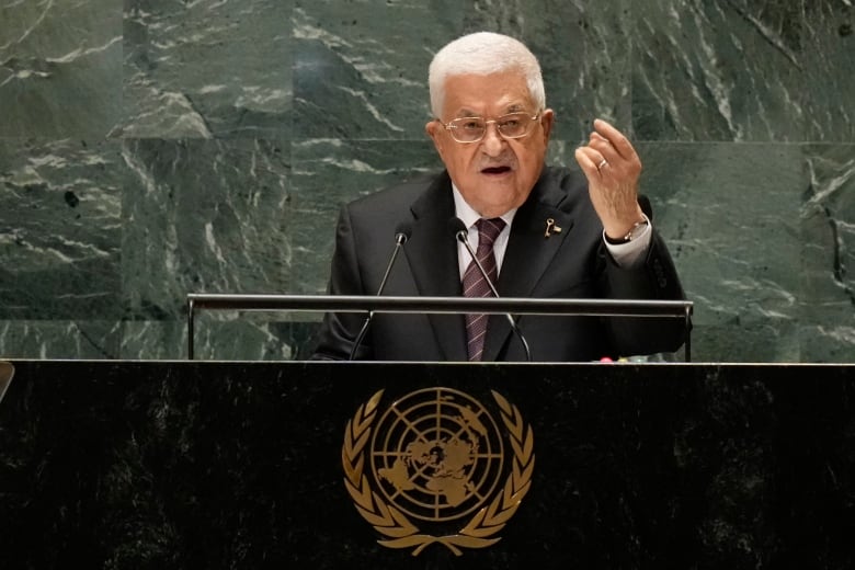 A white-haired man with glasses and facial hair in a suit and tie gestures with his hand while speaking at a podium.