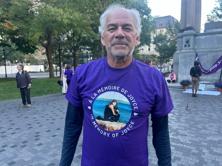 A man with grey hair. He's wearing a purple shirt with the picture of a woman sitting on a rock by a lake. Around the picture are the words: "in memory of Joyce" in English and in French.