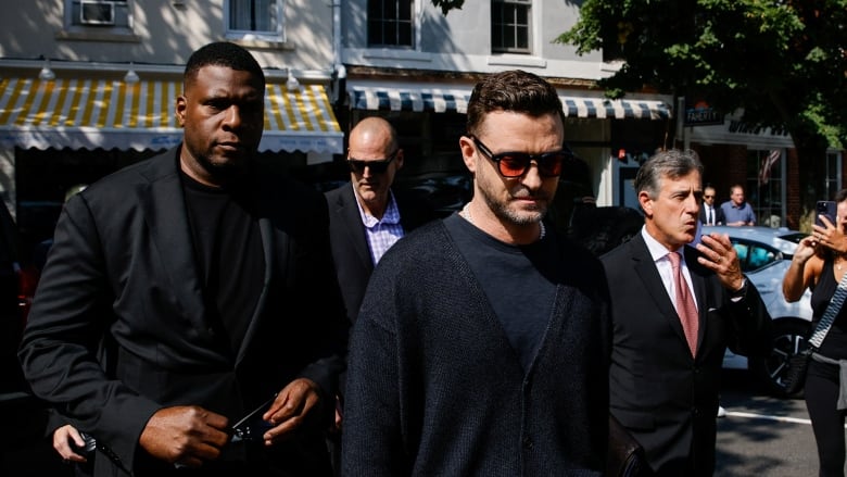 A man in a black sweater and sunglasses with orange lenses walks into court accompanied by a personal security guards.